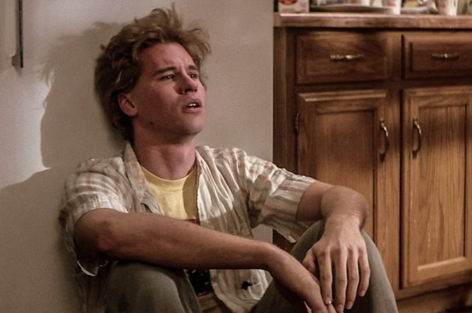 A person with tousled blond hair sits on the floor against a wall, leaning on one knee, looking up.