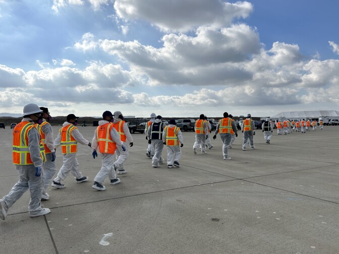 People in white suits and orange safety jackets walk across a large concrete parking lot. 
