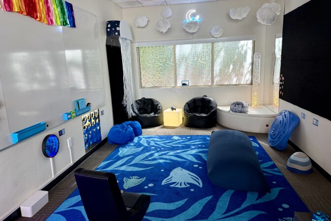A large blue rug with images of kelp and fish covers the floor. There are several black bean bag chairs. The windows are covered with a prismatic film that filters the softens the sunlight. There are puffy white clouds hanging from the ceiling and bubbles fill a long tube-shaped lamp in the corner. 