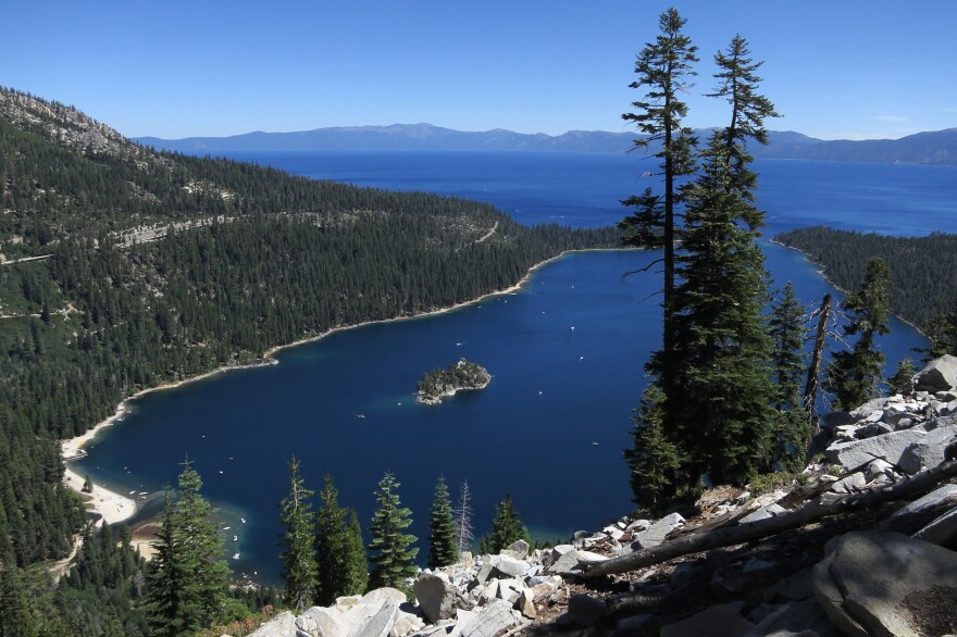 An inlet of deep blue water is dotted with small boats, crystalline lighter blue waters extend beyond the pine tree surrounded bay.