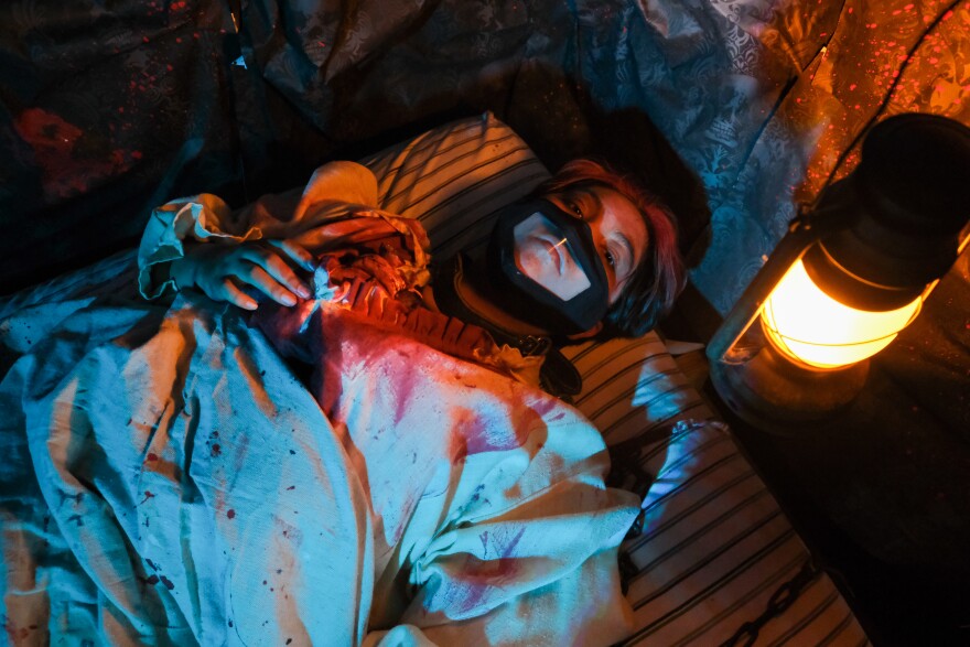 A high school actor lying in a bed in blood-speckled robes portrays a hotel guest with a bloodied chest. The room is dark and a solitary lantern hangs overhead.