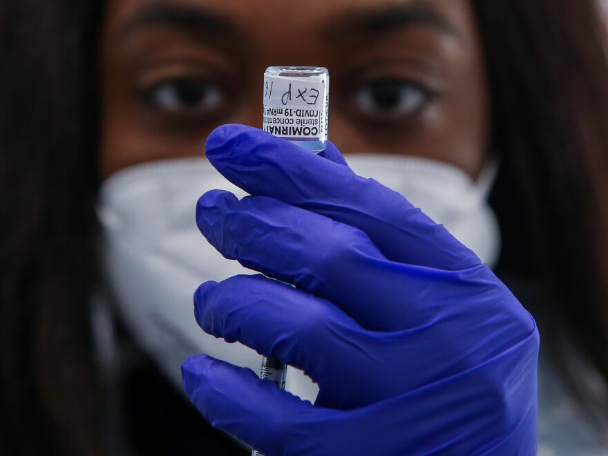 A person in a white mask and blue gloves holds a vaccine vial.