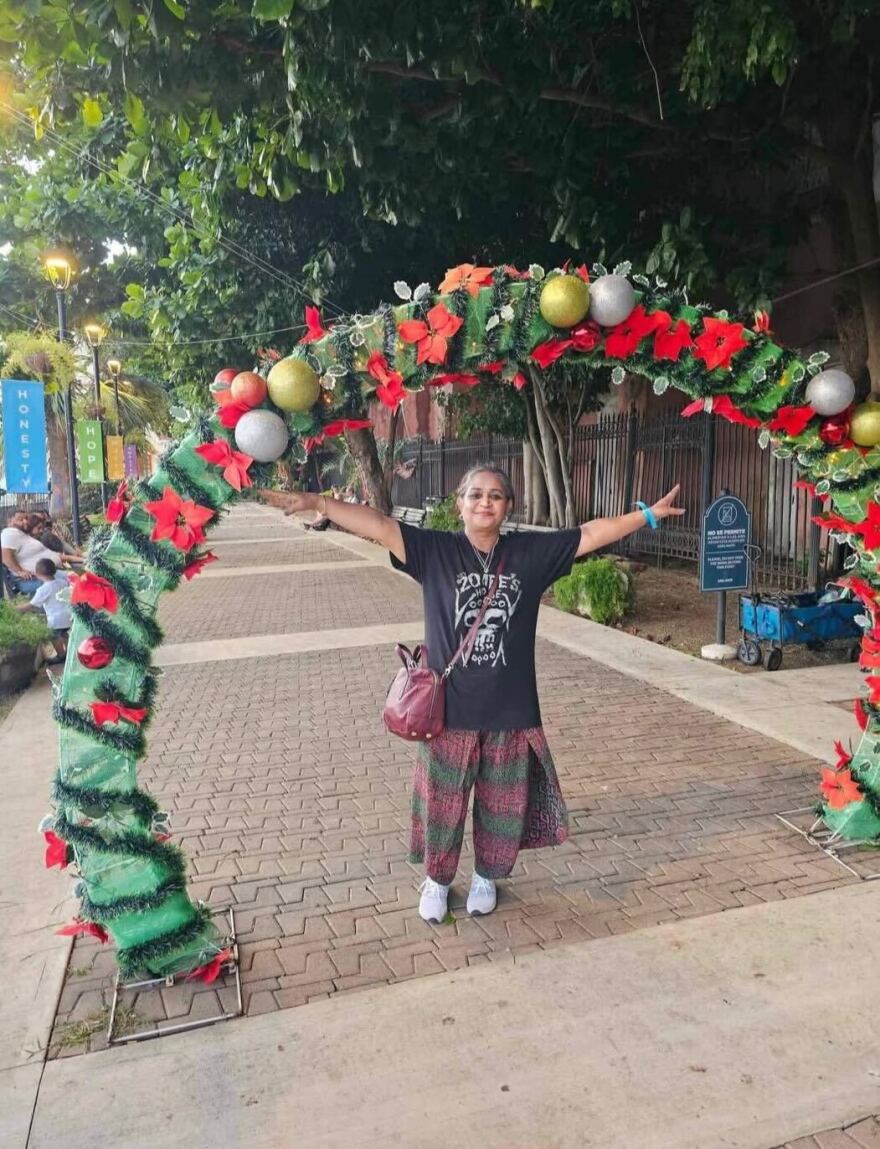 A South Asian woman wearing green and red pants and a black t-shirt and carrying a maroon bag stands under a giant Christmas arc.