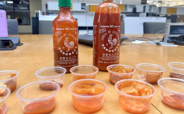 A smaller, mostly empty bottle of Sriracha and a larger, mostly full bottle sit behind two rows of clear used condiment cups.