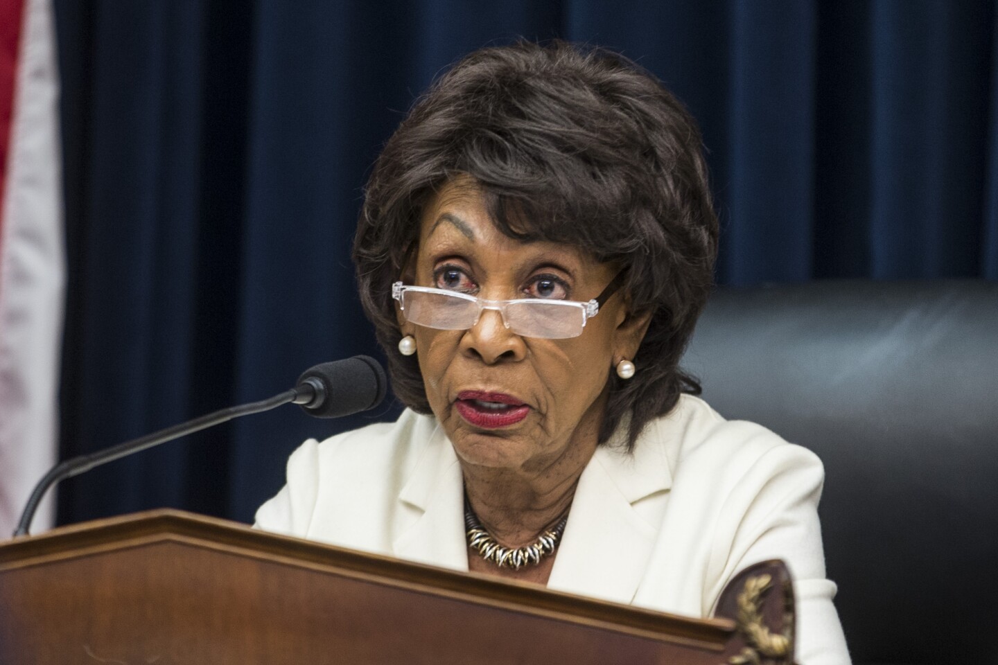 Congresswoman Maxine Waters speaking into a microphone at a lectern while seated in a black chair. She is wearing reading glasses that are halfway down her nose, a necklace, earrings and a white jacket.
