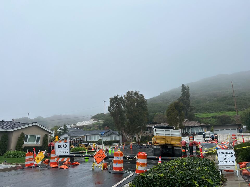 Large orange and white cones and police tape block a road where a landslide has taken place. A hillside covered in a tarp held down by sandbags is visible on the hill behind homes.