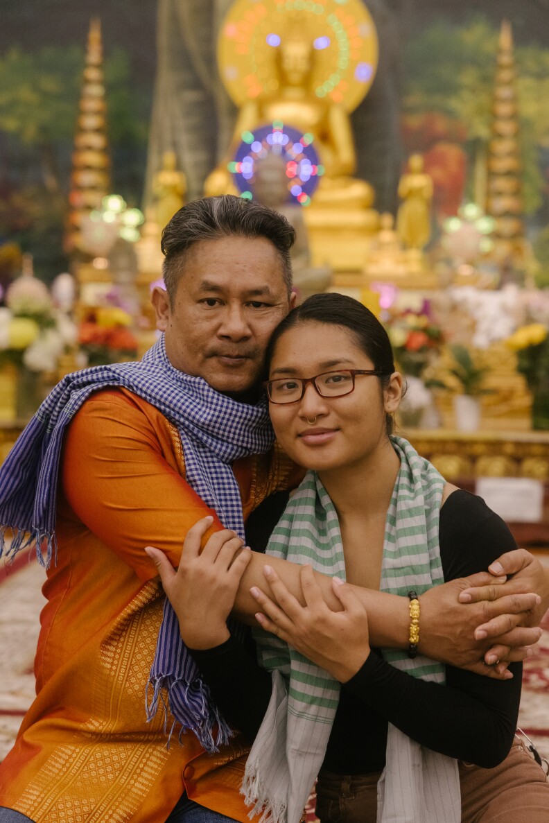 An Asian woman embraces an older Asian man as they stand in front of a gold altar while smiling for a photograph.