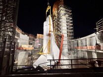 A space shuttle structure towers over the partially-constructed concrete walls and scaffolding that surround it. The structure is displayed vertically towards the pitch black night sky. 