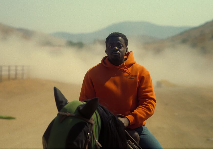 A man with dark skin tone in an orange hoodie sits on a horse in a wilderness landscape, as dust swirls behind him.
