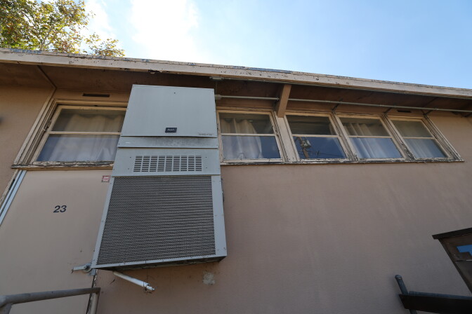 The side of a beige building shown from below.  The frames around the windows are damaged and there is a gray AC unit. 