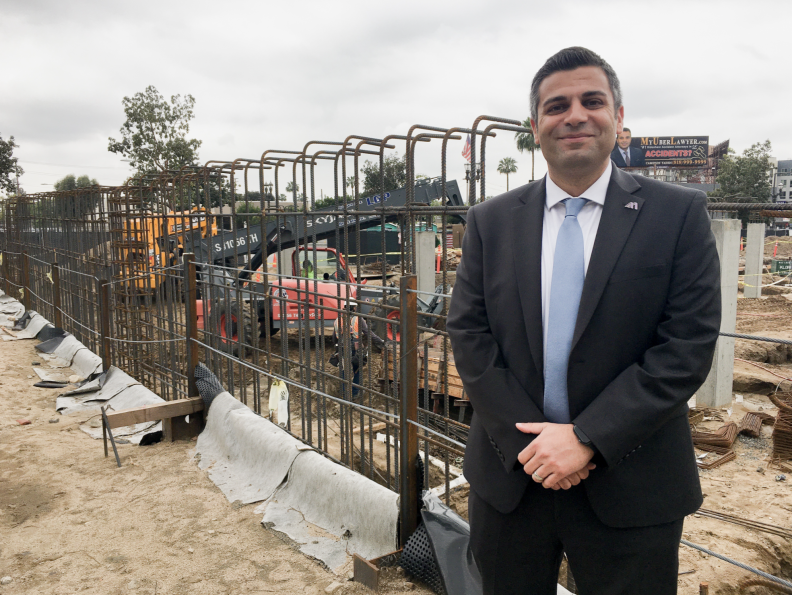 A man in a dark suit and light blue tie stands to the left of a construction site, with rebar and construction vehicles in the background.