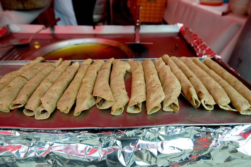 17 corn tortillas are rolled into narrow cylinders with meat visible inside, and aligned on a metal pan. Two spatulas can be seen in the background. Aluminum foil is placed under the pan.