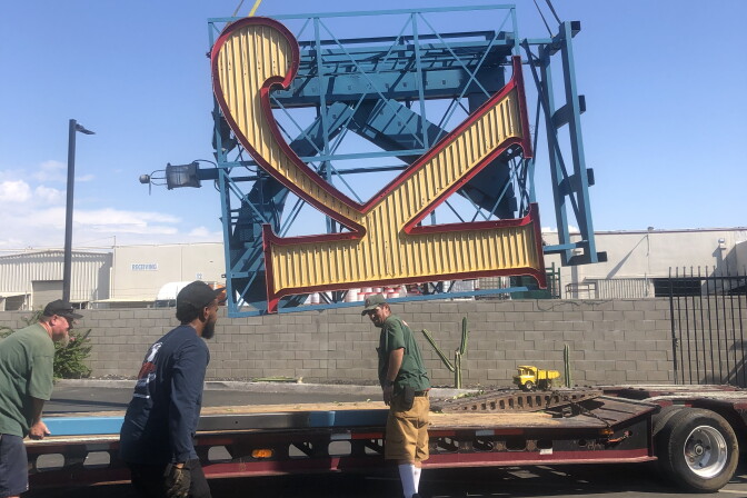 Three workers surround a flatbed truck, as a gigantic sign of the letter "K" -- tilted on its side -- is being lowered onto it