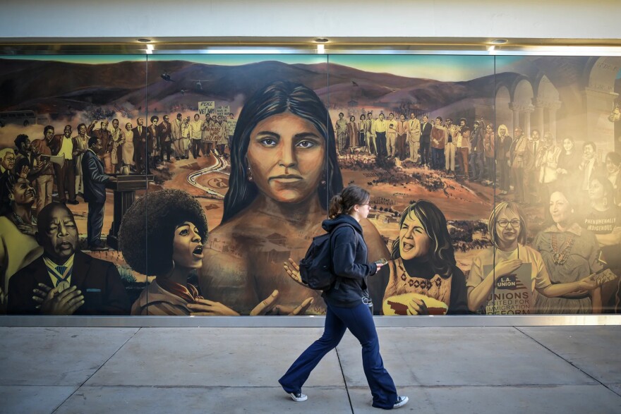 A female brown-haired college student walks by a mural on campus. She is wearing dark jeans, a black sweatshirt, and a black backpack. The mural depicts famous activists in a circle, with a brown-skinned woman with straight dark hair at the center, larger than the rest, and looking at the viewer.