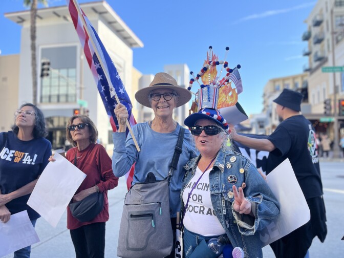 A group of people participating in a street protest or demonstration in an urban setting with modern buildings in the background. One person is wearing a wide-brimmed hat, a blue long-sleeve shirt, and a gray crossbody bag. This person is holding a large American flag on a wooden pole. Another person is wearing a denim jacket adorned with multiple pins and buttons, along with a white shirt that reads “DANCING FOR DEMOCRACY.” 