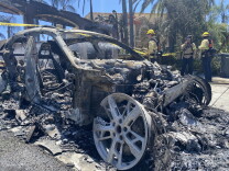 The burned out husk of a car, wheels left with no tire around them, ashen pieces/remnants all around, as firefighters stand nearby. Yellow caution tape extends in the background, with an intact house behind as well.