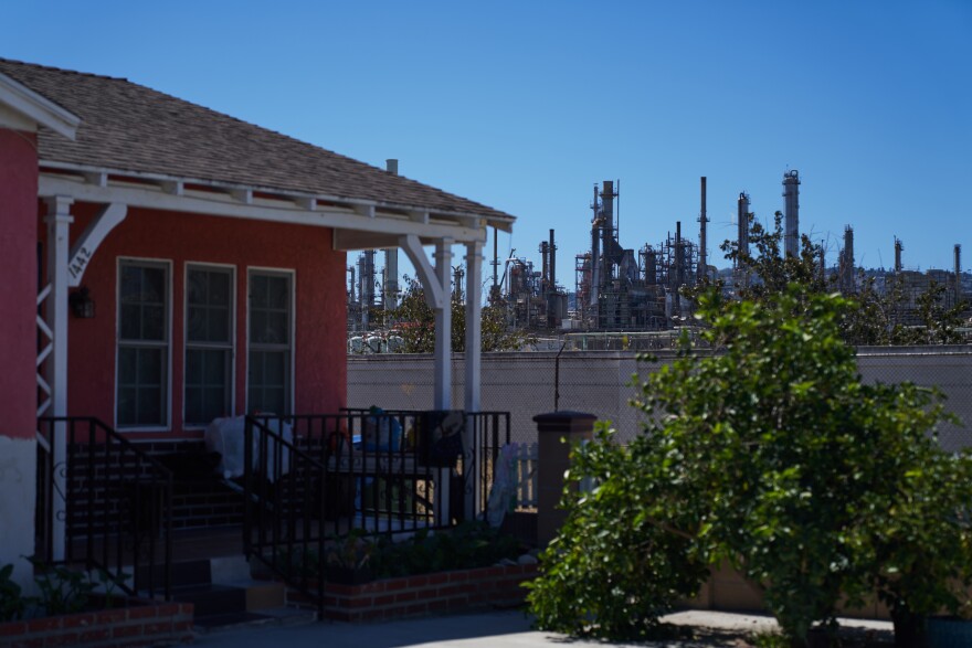 A home with red walls and a slate roof with chairs on a small porch with a large oil refinery in the background
