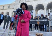BLM co-founder Patrisse Cullors speaks into several microphones outside LA city hall. A banner behind her reads "Black Lives Matter." City council members and several activists stand behind her. Cullors wears a bright pink coat. 