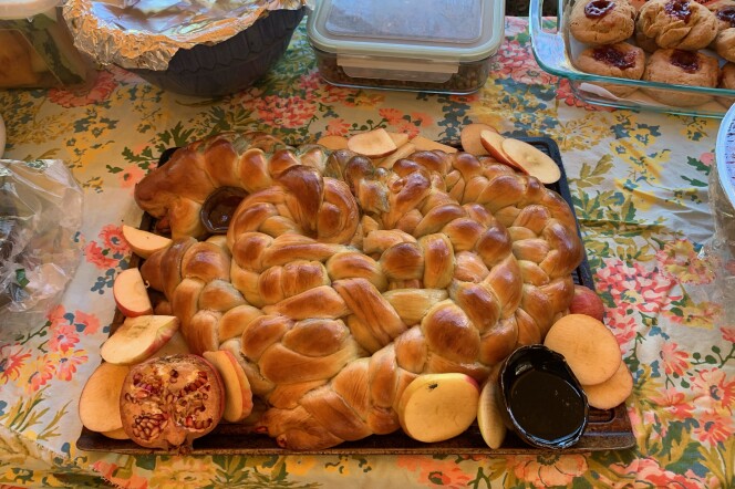 A braided loaf of challah bread served on a platter with apples, pomegranates and honey, on top of a floral tablecloth.