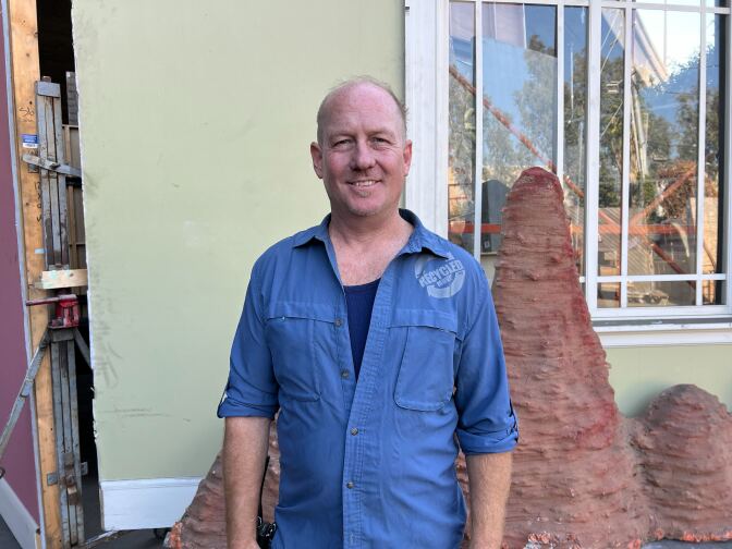 A middle aged white man in blue coveralls smiles for the camera in front a structure that looks small mountains