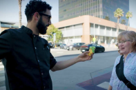 A man with a light skin tone and dark hair, wearing glasses and a black chef's coat, extends a colorful taco toward a woman with light hair and a light skin tone on a sunlit Los Angeles sidewalk. The woman, wearing a plaid shirt with a crossbody bag, smiles mid-conversation as office buildings and palm trees line the street behind them.