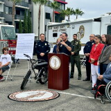A man speaks at an Orange County District Attorney podium beside an e-bike, with officials and vehicles from fire, sheriff and DA departments behind him.