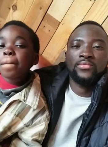 An older Black man with black hair and facial hair takes a selfie with a younger Black kid. The man wears a white t-shirt with a black jacket and the young boy wears a red t-shirt with a brown flannel over it.