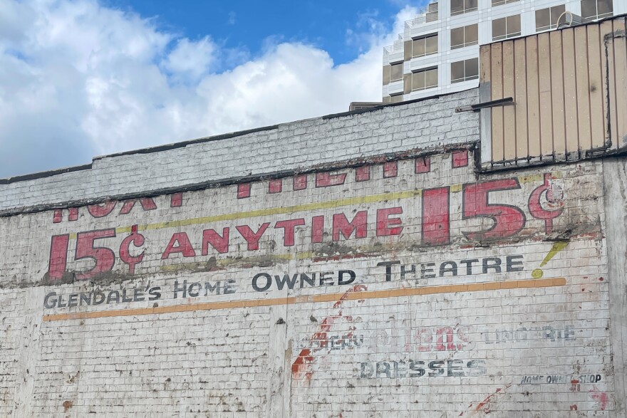 Faded paint on the side of a brick building reads "ROXY THEATRE; 15 CENT ANYTIME; GLENDALE'S HOME OWNED THEATER." Some lower faded type reads "Dresses" and "Lingerie." A blue sky is seen in the background, a more modern building to the right.