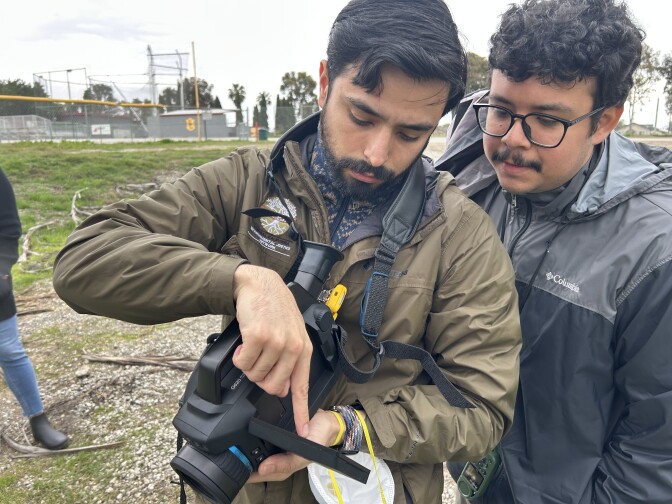 Two young men with light brown skin land dark hair look at the viewfinder of a FLIR infrared video camera.