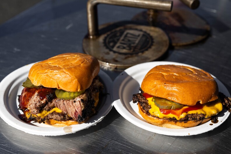The scene is a food pop-up, where a metal table sits beneath a tent. Atop the table are two paper plates holding burgers. The burger on the left is a brisket burger -- there are juicy slices of brisket sliding out. On the right, a traditional American cheese smashburger. In the background are two burger presses used to smash the burgers thin. The heavily-weighted presses have the name "Bartz" emblazoned on the background, in a touch of branding.