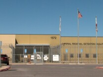 A one story, brown building behind a barbed wire fence. Three flagpoles are in front of the building. The middle flagpole is flying the American flag, the two white, unfurled flags hang on the other two poles. On the building is signage that reads, "Imperial Regional Detention Facility."