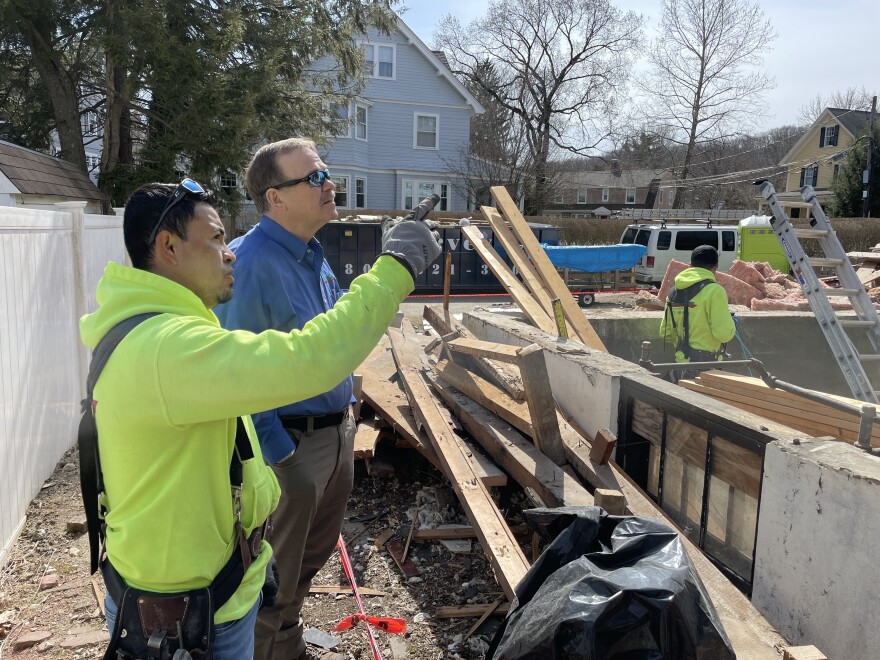 A man in a lime green hoodie point with a gloved hand at a construction site while standing next to another man wearing a long-sleeved button down shirt.