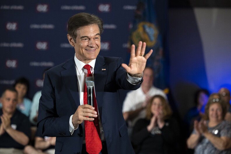 Mehmet Oz, a tan celebrity physician and Republican Senate candidate, wears a blue suit with a white shirt and a red tie and holds his left hand up waving while holding a microphone, gray and black, close to his chest while smiling in a room where several people applaud in the background.