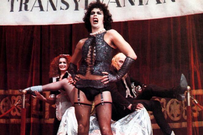 Tim Curry as Dr. Frank-N-Furter in The Rocky Horror Picture Show. He wears curly black hair, a white pearl necklace, a sparkly black vest, black panties, black gloves, and black thigh-high stockings.