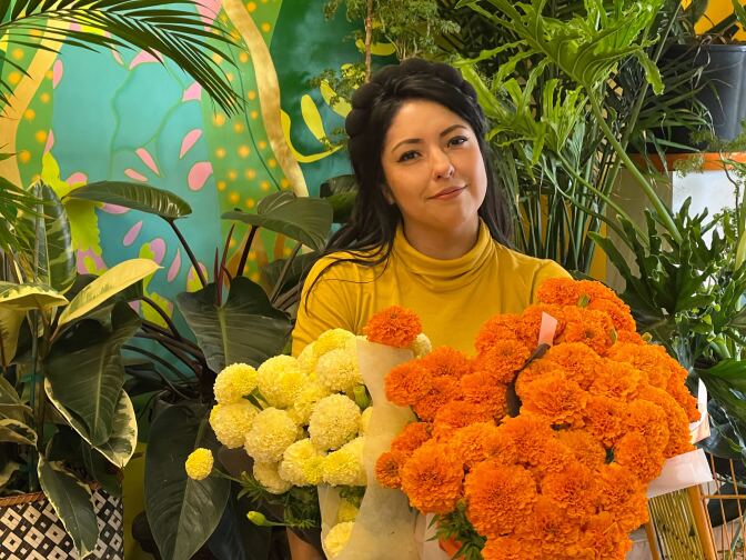 a Latina wearing a yellow turtleneck smiles holding big bunches of orange and yellow marigolds in front of a collection of green plants in a florist shop