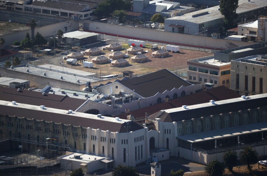 The gothic outer rim of San Quentin prison is seen in the foreground. In the background, tents erected to care for COVID-19 patient are in an open yard.