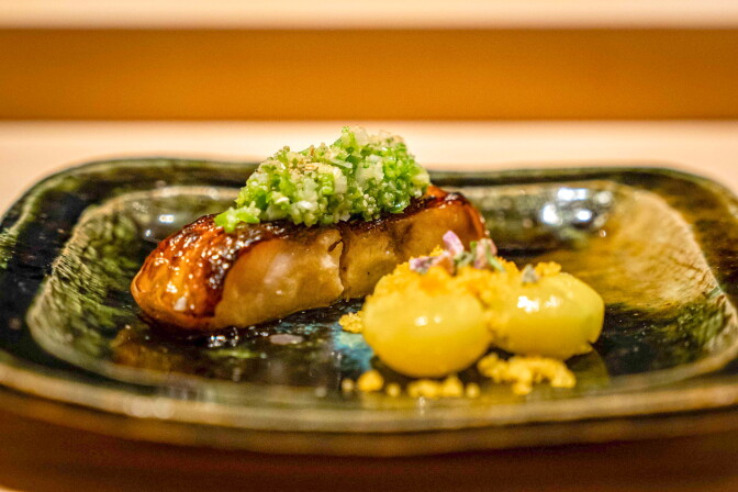 A mottled green glazed dish holds a piece of fish with a relish on top, and two yellow round balls which have been opened to see the inside.