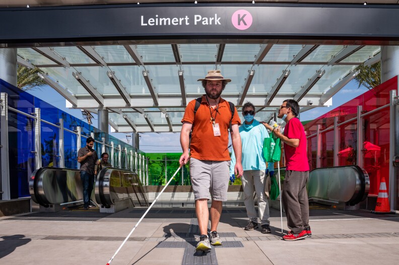 A man walks along a textured pathway using a long white cane for the visually impaired to guide him. He's wearing a dark orange polo shirt, gray shorts and a sun hat, and he's passing beneath a sign that reads "Leimert Park" followed by a letter K in a pink circle, for the Metro K Line.