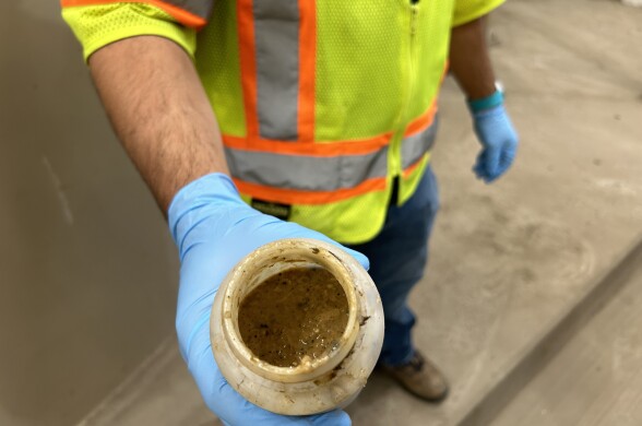 A worker wearing blue gloves and a safety vest and jeans holds a water bottle-sized container of food waste slurry. 