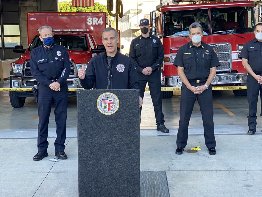 LA Mayor Eric Garcetti speaks at a podium, flanked to our left by LAPD Chief Michel Moore and to our right LA Fire Chief Ralph Terrazas. Two other fire department staff stand behind Terrazas, and a fire engine and ambulance are behind them.