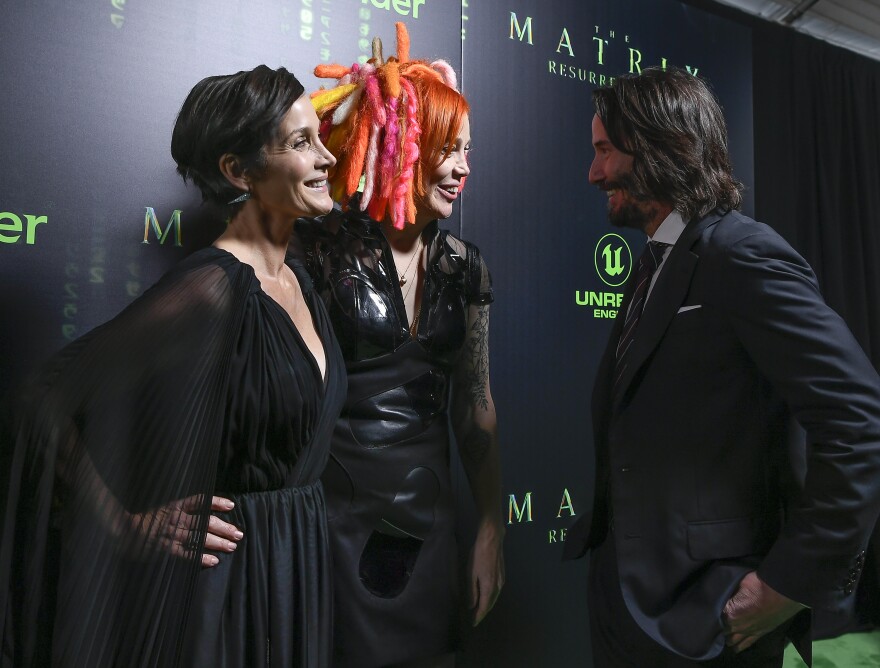 Three people talk in front of a dark background with bright green lettering touting "The Matrix Resurrections."