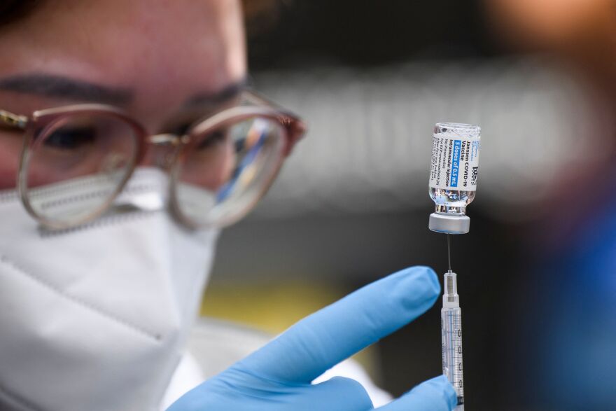 A needle and vial of COVID-19 vaccine is in focus as a masked healthcare worker wearing glasses touches the needle with a gloved finger.