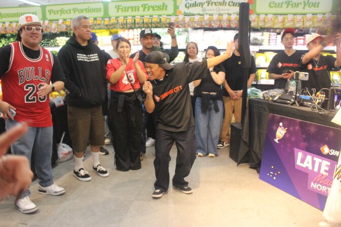 A person in a Seafood City shirt dances with arms outstretched inside a Seafood City supermarket as a crowd watches and cheers.