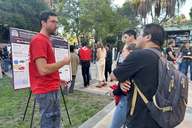 A man with light skin tone stands before a scientific research poster in a courtyard. In front of him, three other people listen as he speaks. In the background, people pause to look at other posters. 