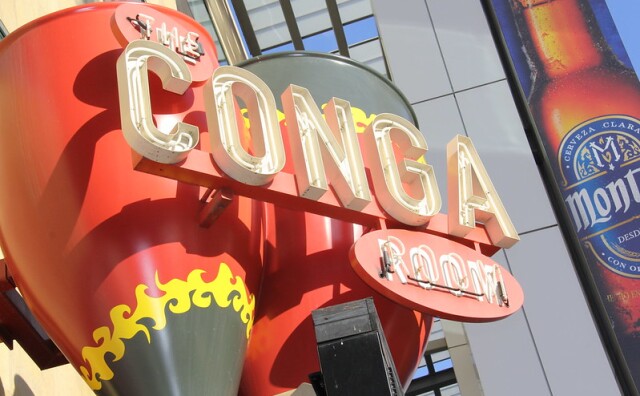 A view looking up at the large sign for The Conga Room at LA Live with the name of the nightclub written in all capital white letters that light up at night, set against red and gray conga drums, trimmed at the bottom with what looks like yellow flames.