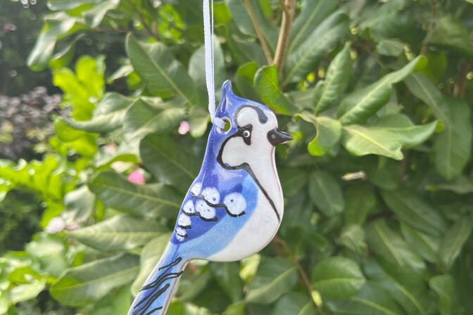 A hand crafted ceramic ornament depicting a blue jay hangs in a tree. 