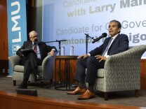 Two men sit on upholstered chairs in suits with mics in front of them. They're on a stage.