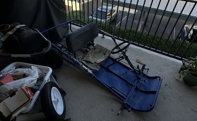 An old, beat up go-kart frame that's missing a few pieces is sitting on an apartment patio overlooking a parking lot.