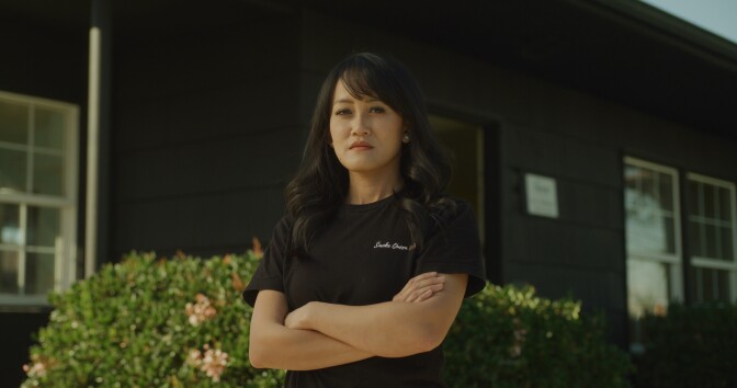 A women with light  brown skin stands in an outdoor setting with her arms crossed and is wearing a black chef's coat
