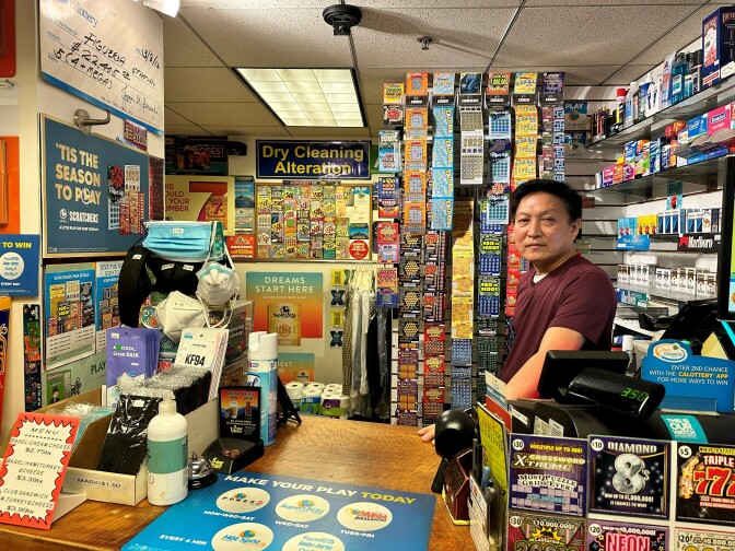 An Asian man stands behind a counter with a cash register. All around and on the walls are small colorful lottery cards, and a shelf behind him displays packs of cigarettes and various medicines. One sign on the wall reads "'Tis the season to play" and another reads "Dry cleaning alterations."